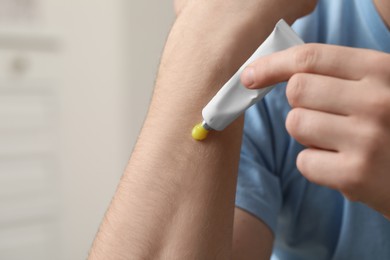 Man applying yellow ointment from tube onto his arm indoors, closeup