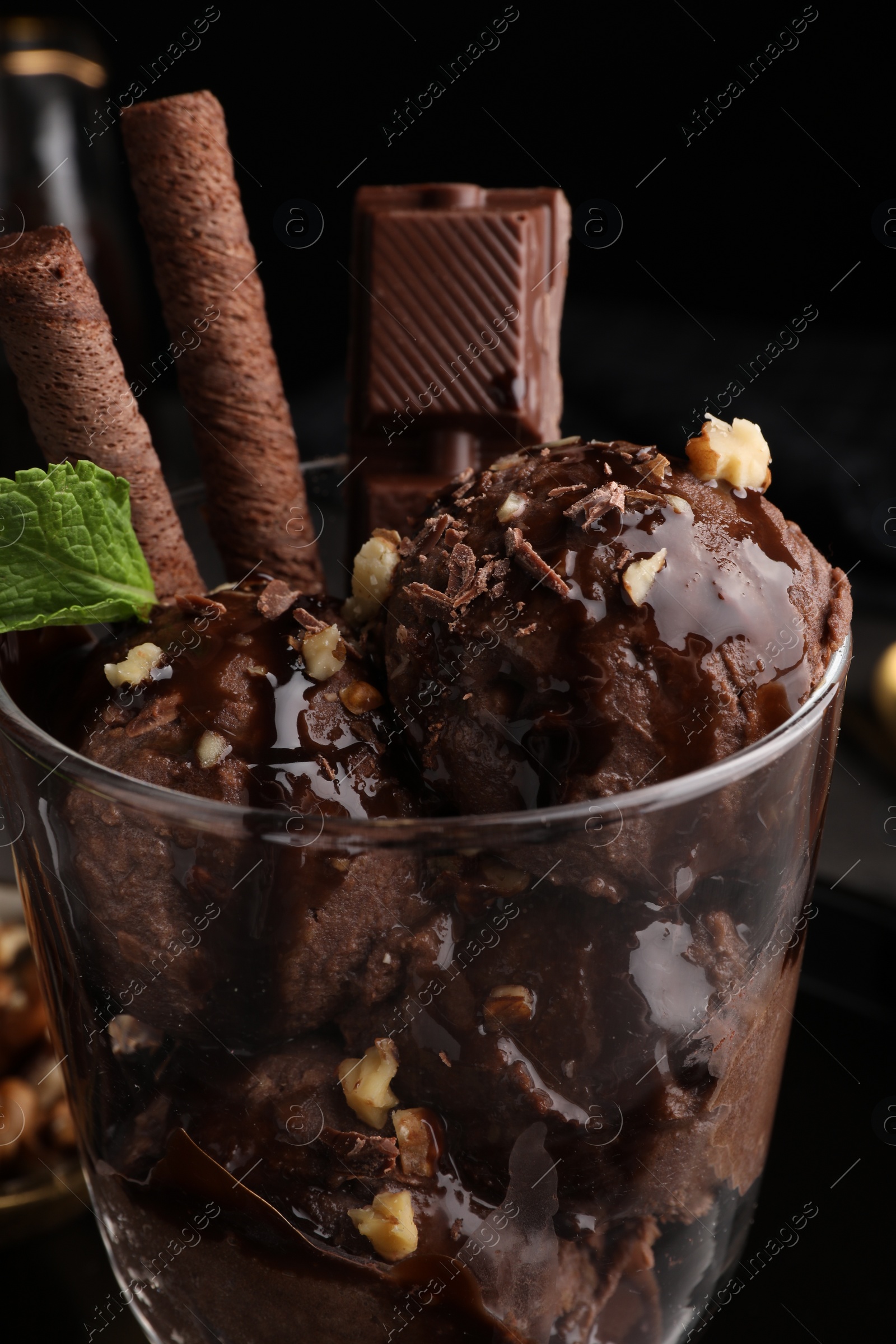 Photo of Tasty chocolate ice cream with sauce, nuts and wafer rolls in glass dessert bowl on table, closeup