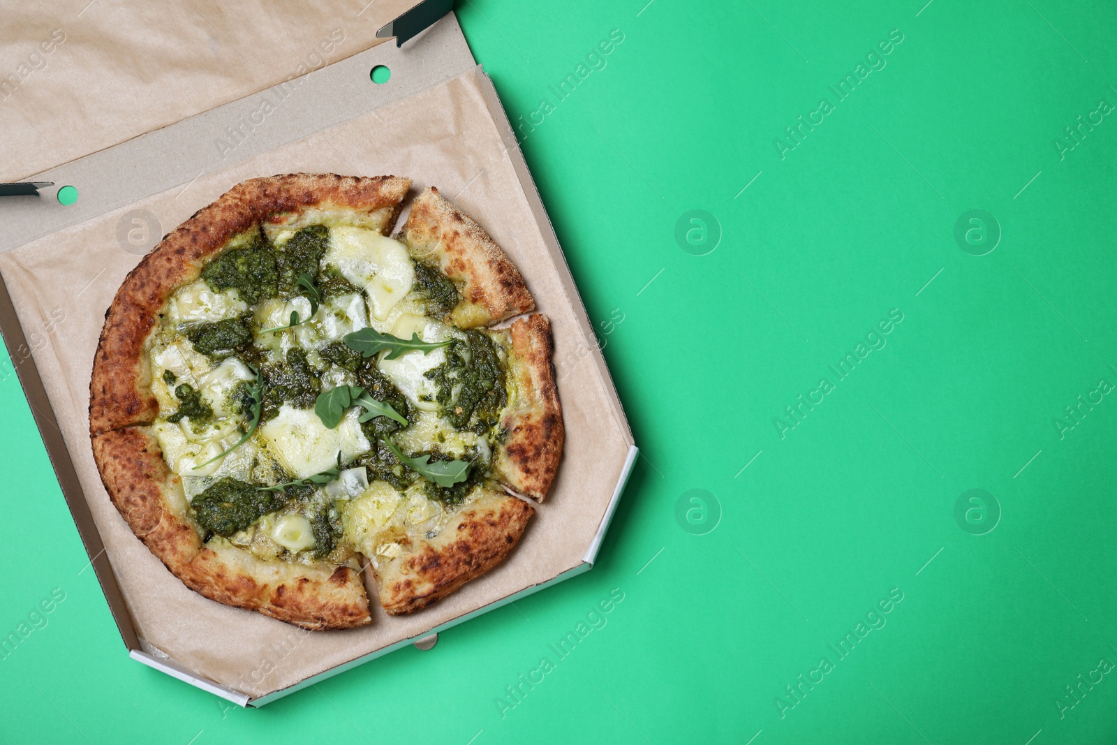 Photo of Delicious pizza with pesto, cheese and arugula in cardboard box on green background, top view. Space for text