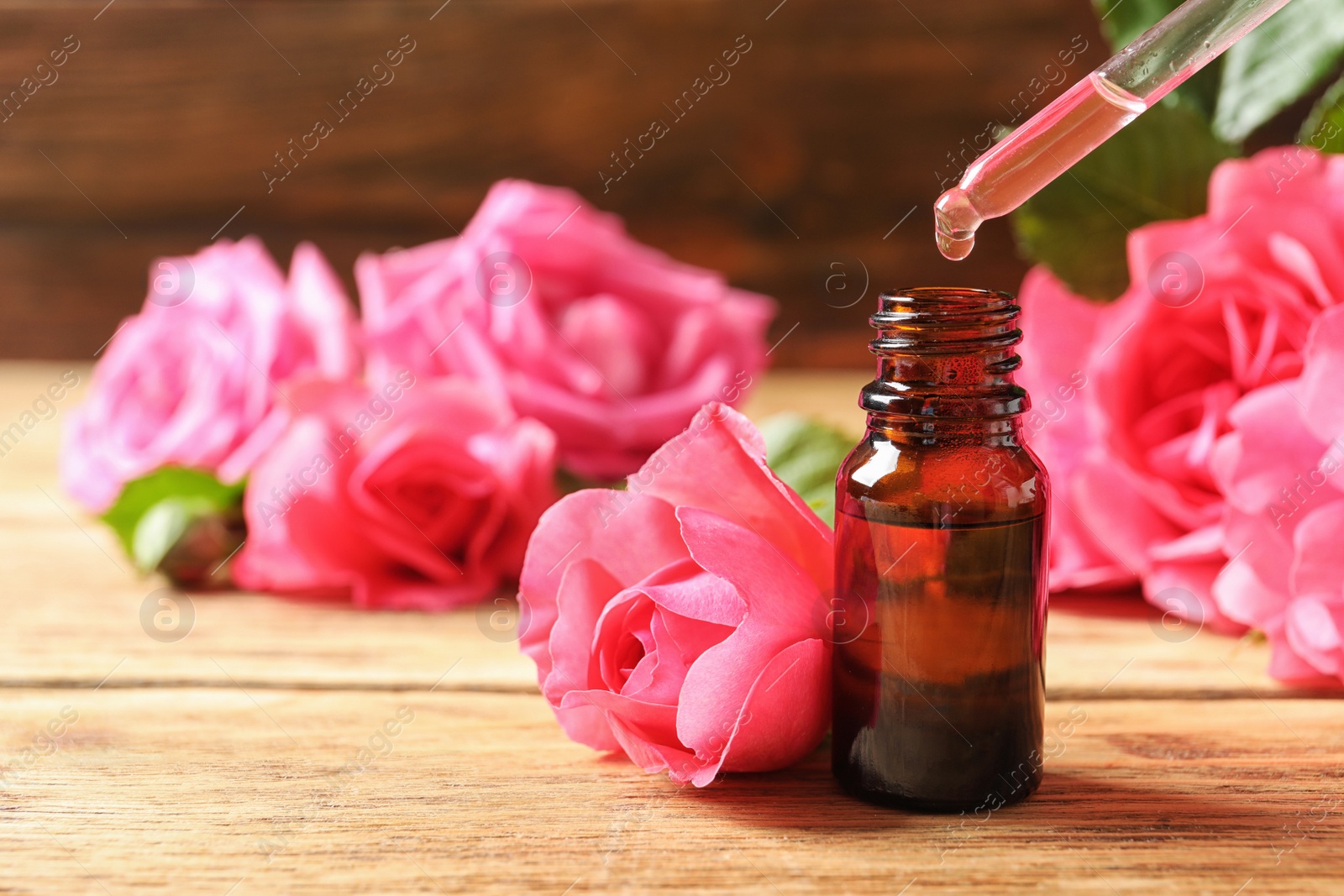 Photo of Bottle of rose essential oil, pipette and flowers on wooden table, space for text