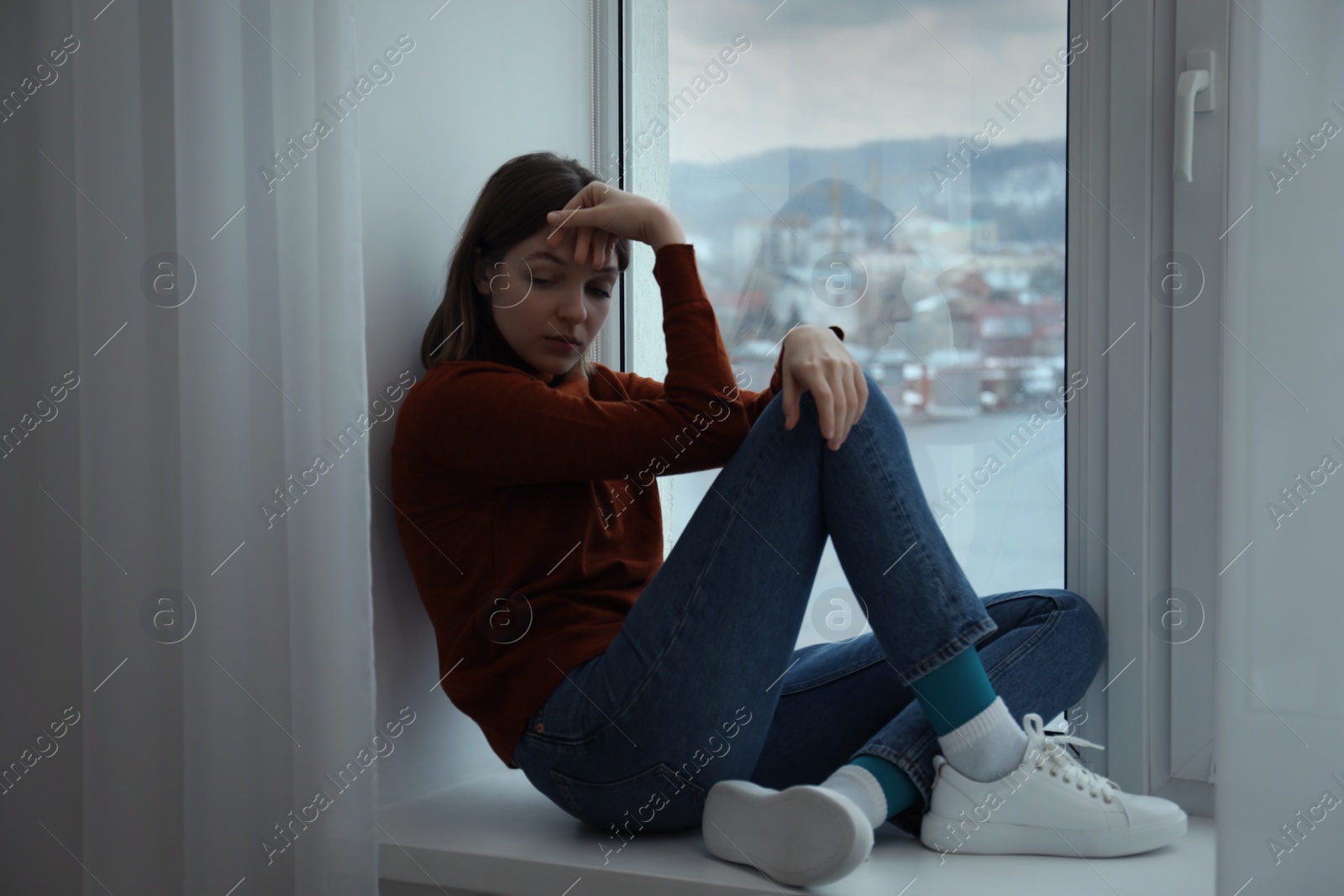 Photo of Sad young woman sitting on windowsill near window at home