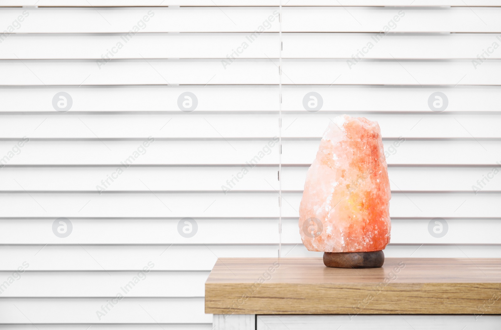 Photo of Himalayan salt lamp on wooden table near window blinds. Space for text