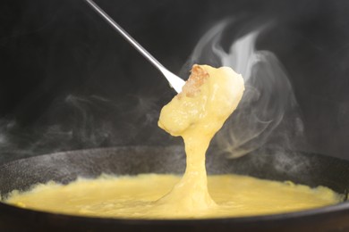 Image of Dipping piece of bread into fondue pot with tasty melted cheese against dark background, closeup