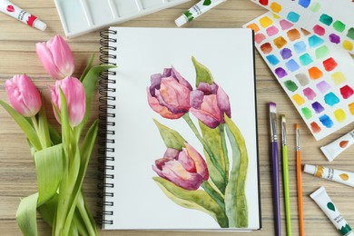 Photo of Painting of tulips in sketchbook, flowers and art supplies on wooden table, flat lay