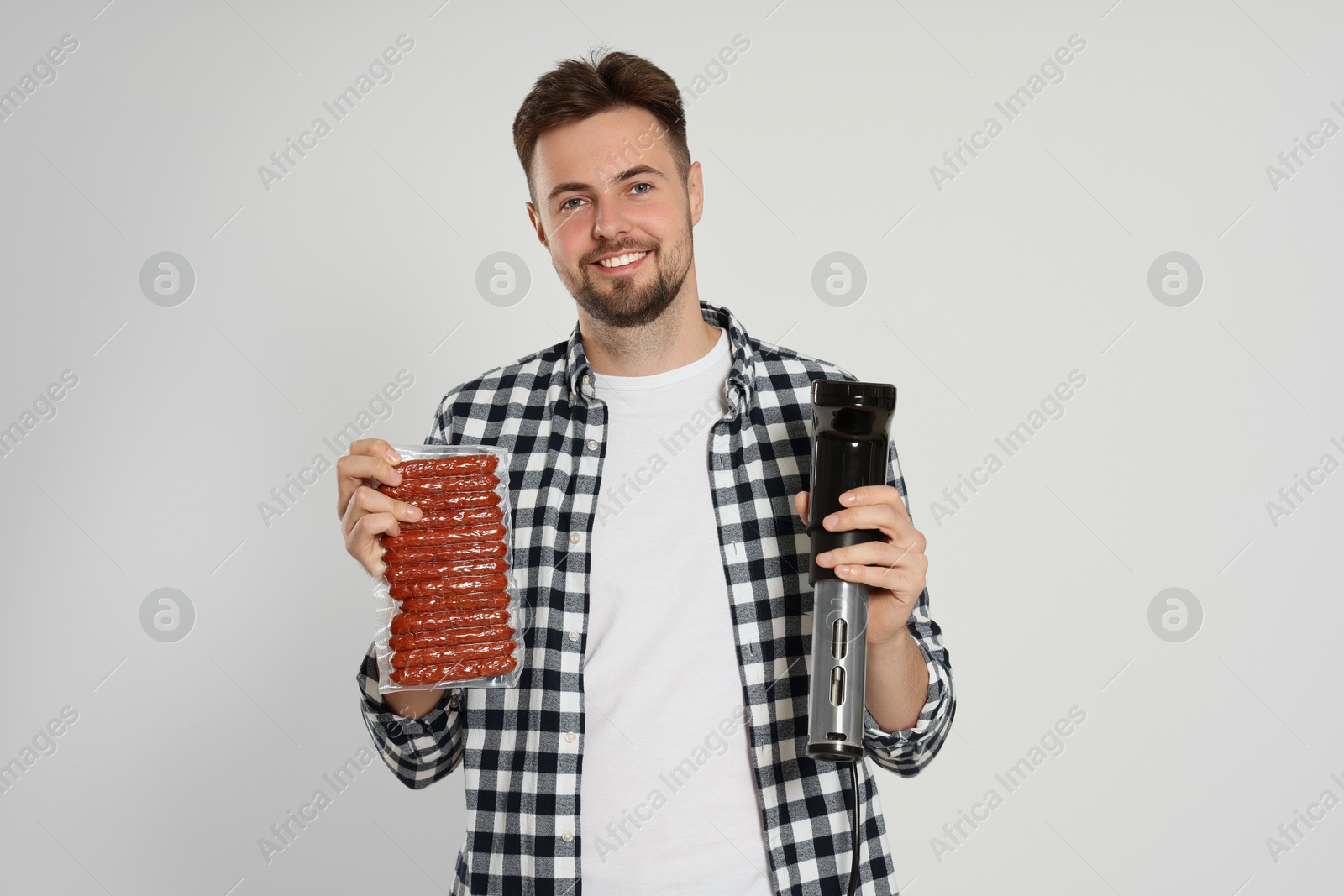 Photo of Smiling man holding sous vide cooker and sausages in vacuum pack on beige background