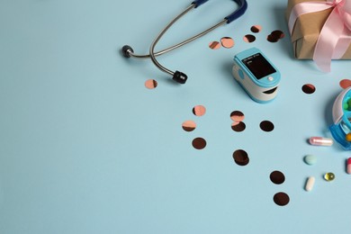 Stethoscope, pills, box and fingertip pulse oximeter on light blue background, space for text. Medical gift