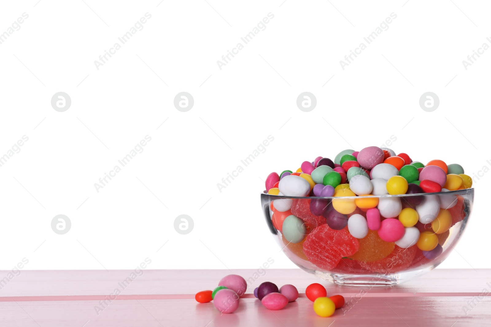 Photo of Many different candies in glass bowl on pink wooden table against white background
