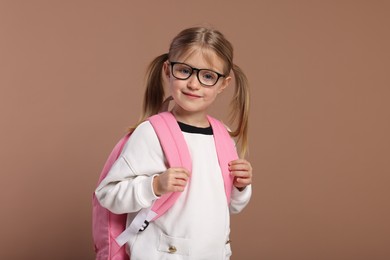 Happy schoolgirl in glasses with backpack on brown background