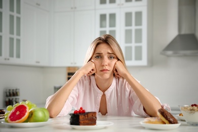 Photo of Woman choosing between sweets and healthy food at white table in kitchen