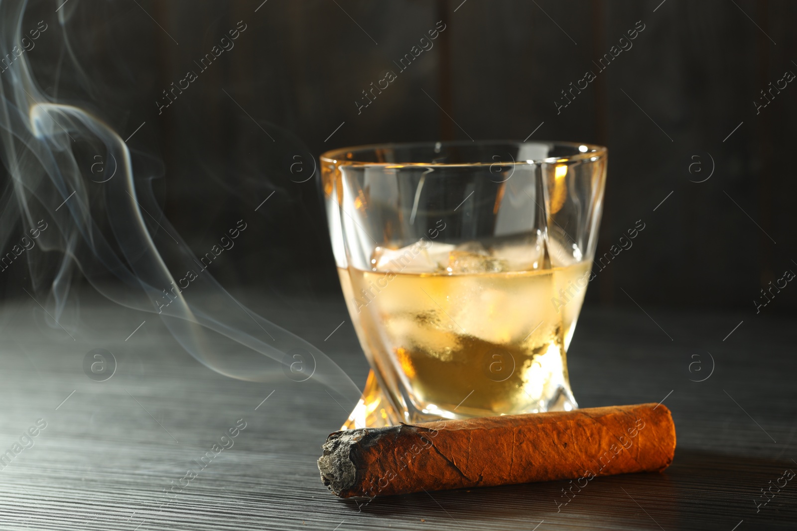 Photo of Glass of whiskey with ice cubes and smoldering cigar on black wooden table, closeup. Space for text