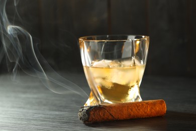 Photo of Glass of whiskey with ice cubes and smoldering cigar on black wooden table, closeup. Space for text