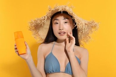 Beautiful young woman in straw hat holding sun protection cream on orange background