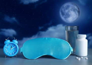 Alarm clock, soporific pills, sleeping mask and glass of water on light grey table against night sky with full moon. Insomnia