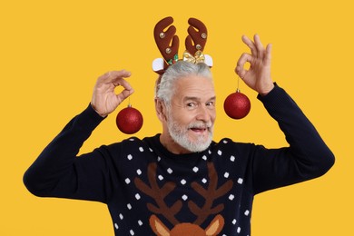 Photo of Senior man in Christmas sweater and reindeer headband holding festive baubles on orange background