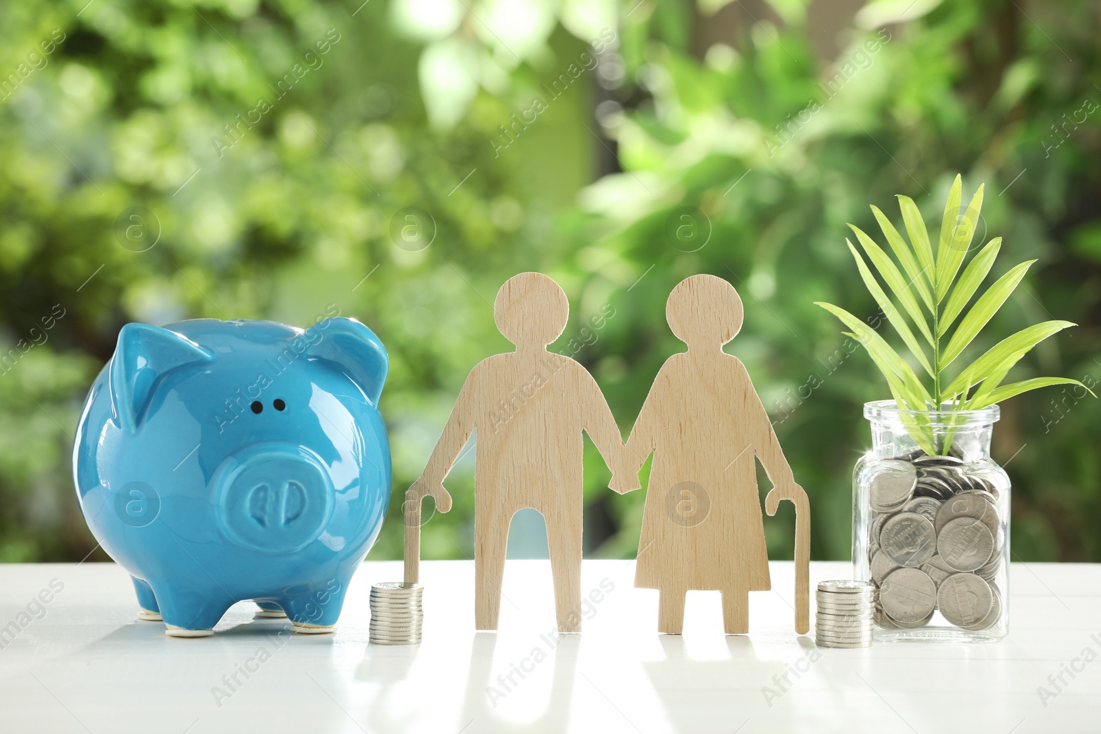 Photo of Pension savings. Figure of senior couple, piggy bank, jar with coins and twig on white table against blurred green background