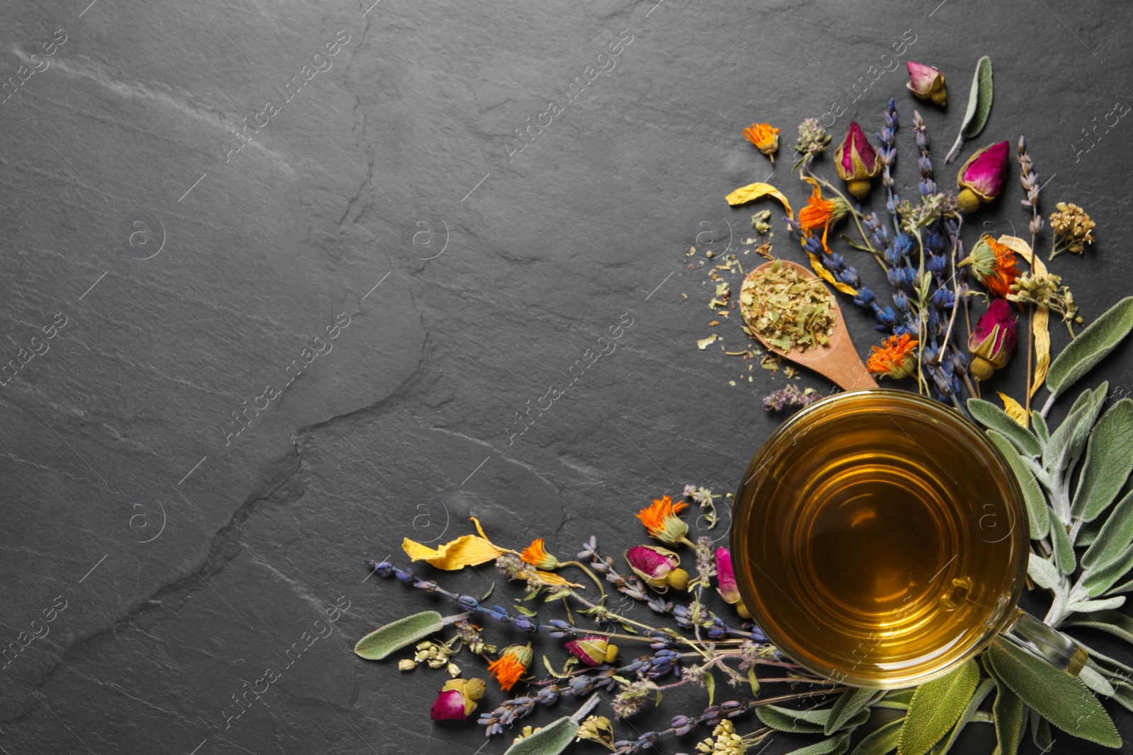 Photo of Glass cup of aromatic freshly brewed tea near different dry herbs on black table, flat lay. Space for text