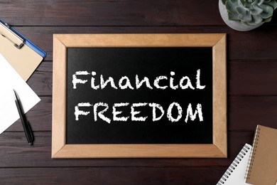 Image of Small chalkboard with text Financial Freedom and stationery on wooden desk, flat lay