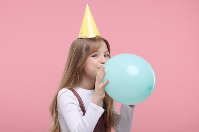 Photo of Cute little girl in party hat inflating balloon on pink background