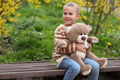 Little girl with teddy bear on wooden bench outdoors. Space for text
