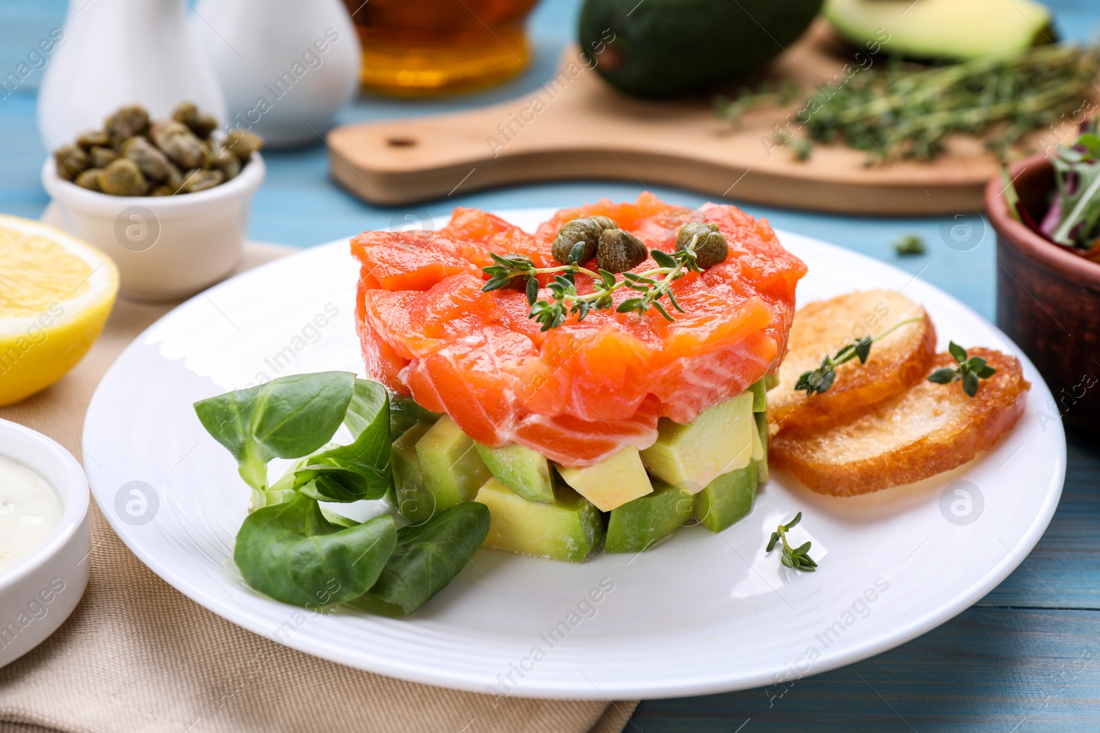 Photo of Delicious salmon tartare served with avocado and croutons on light blue table, closeup