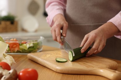 Photo of Woman cutting cucumber at wooden table in kitchen, closeup