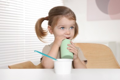 Cute little child with bottle and cup of tasty yogurt at white table indoors