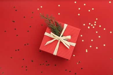 Photo of Christmas gift box with coniferous twigs and shiny confetti on red background, flat lay. Space for text