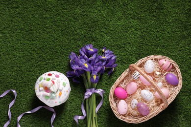 Photo of Easter basket with painted eggs, iris flowers and tasty cake on green grass, flat lay. Space for text
