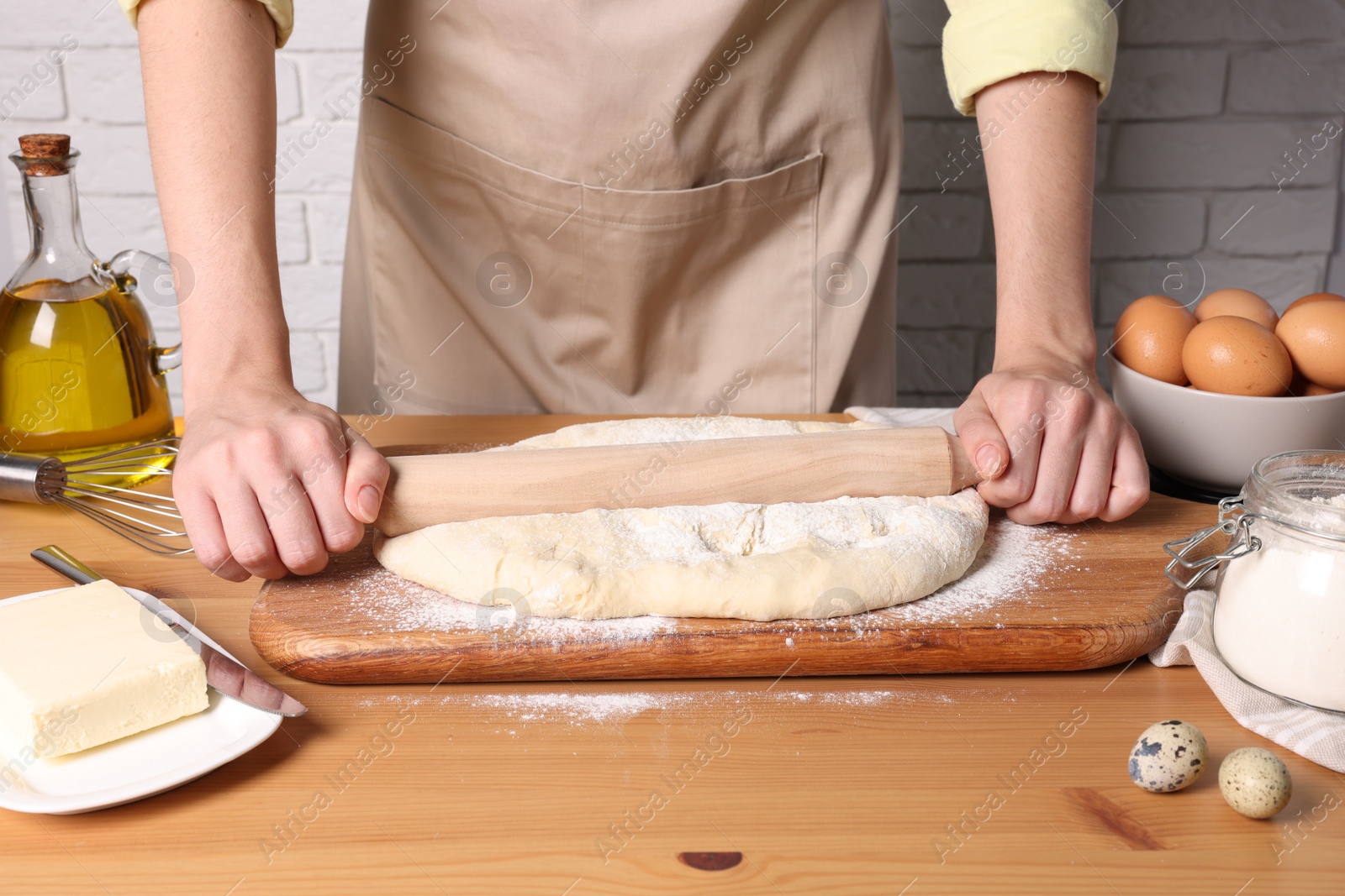 Photo of Woman rolling dough with wooden pin at table near white brick wall, closeup