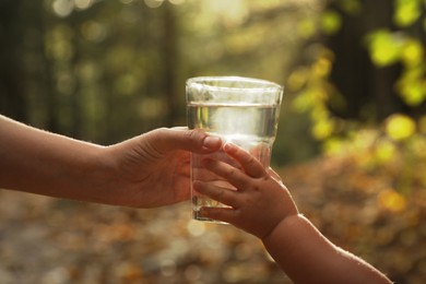 Photo of Mother giving her daughter glass of fresh water in forest on sunny day, closeup
