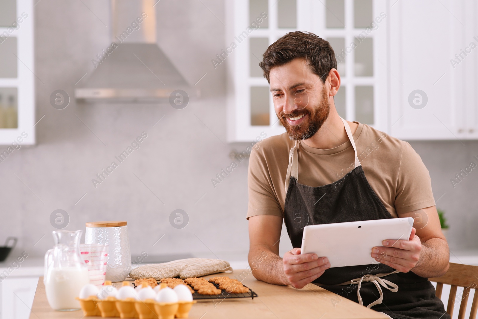 Photo of Man with freshly baked cookies watching online cooking course via tablet in kitchen