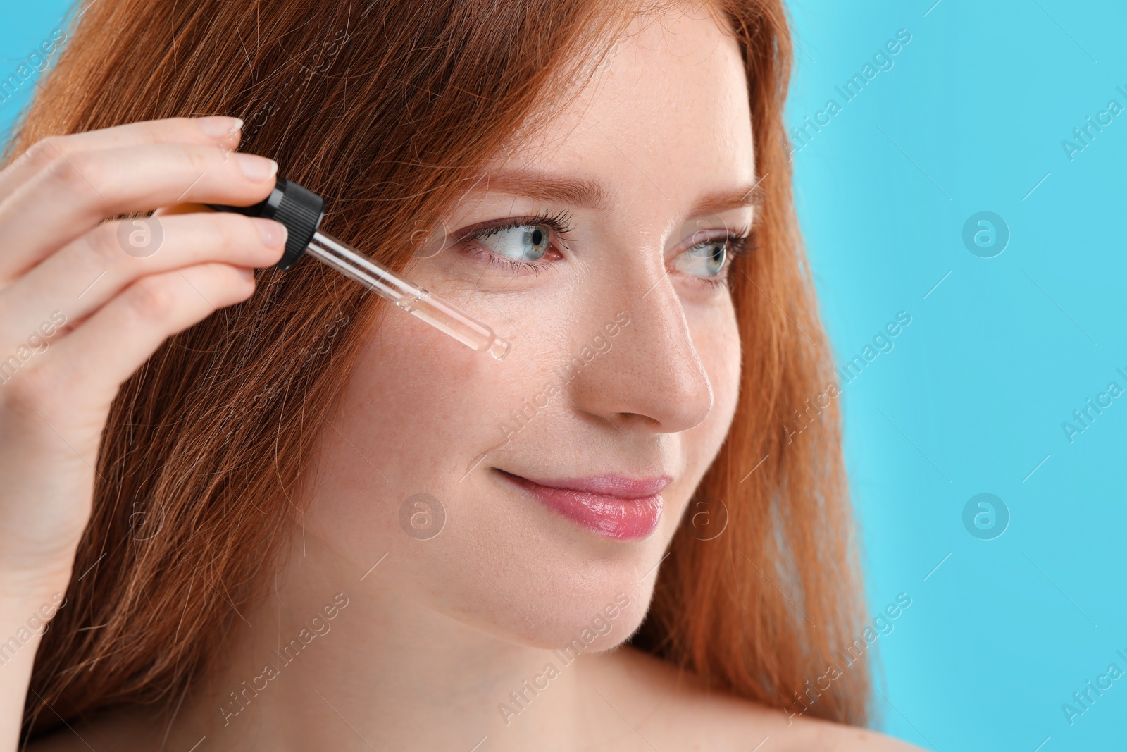 Photo of Beautiful woman with freckles applying cosmetic serum onto her face against light blue background, closeup