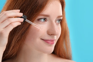 Beautiful woman with freckles applying cosmetic serum onto her face against light blue background, closeup