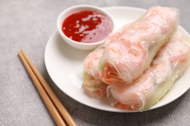 Photo of Tasty spring rolls with sauce and chopsticks on grey textured table, closeup