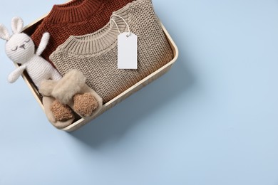 Photo of Different baby accessories, knitted sweaters and blank card in box on light blue background, top view. Space for text