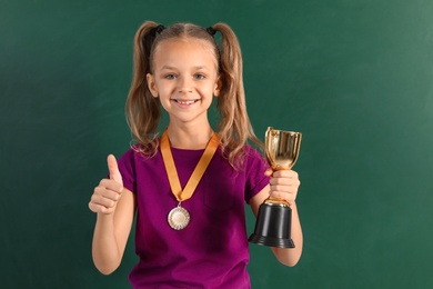 Photo of Happy girl with golden winning cup and medal near chalkboard