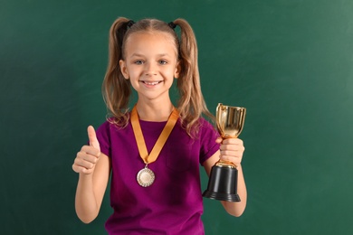 Happy girl with golden winning cup and medal near chalkboard