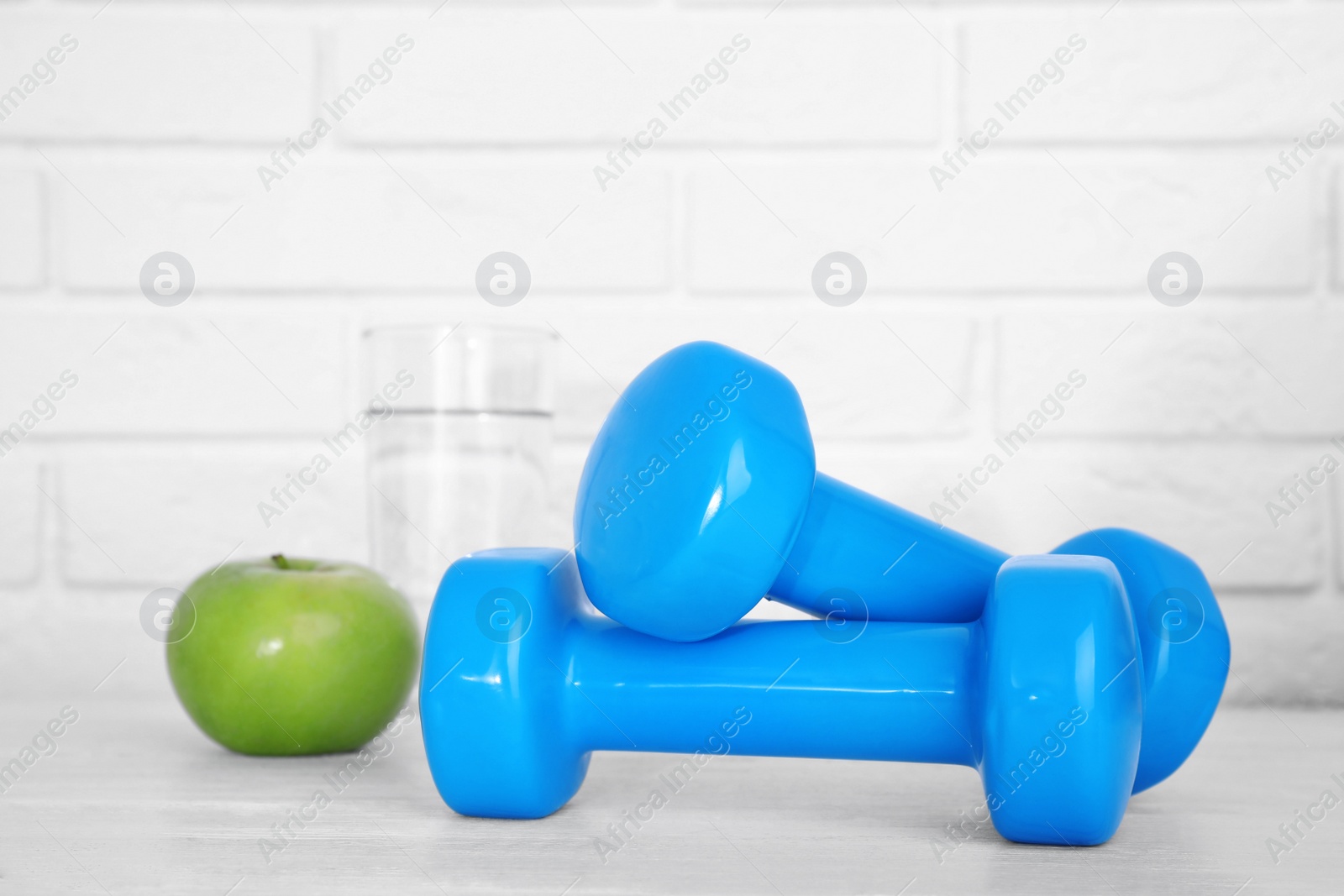 Photo of Stylish dumbbells, apple and glass of water on table against brick wall, space for text. Home fitness