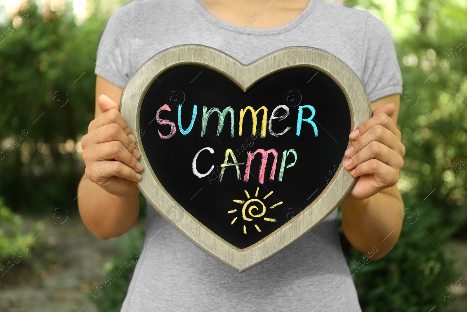 Photo of Woman holding heart shaped blackboard with text SUMMER CAMP and drawing on blurred background, closeup