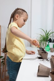 Photo of Little girl adding soil into peat pots on window sill indoors. Growing vegetable seeds