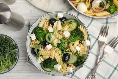 Photo of Bowl of delicious pasta with cucumber, olives, broccoli and cheese on white wooden table, flat lay