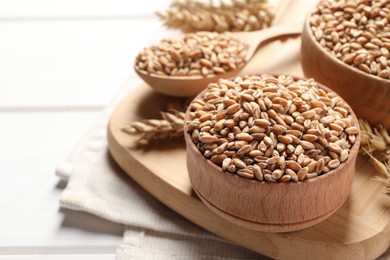 Wheat grains in bowl and spoon on wooden board, closeup view