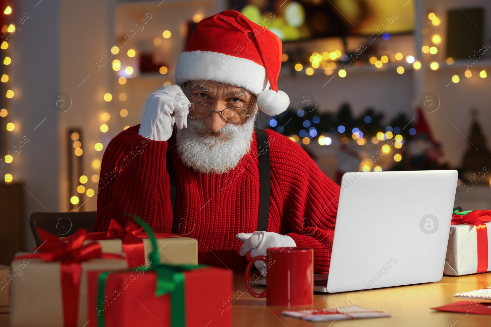 Photo of Santa Claus using laptop at his workplace in room decorated for Christmas