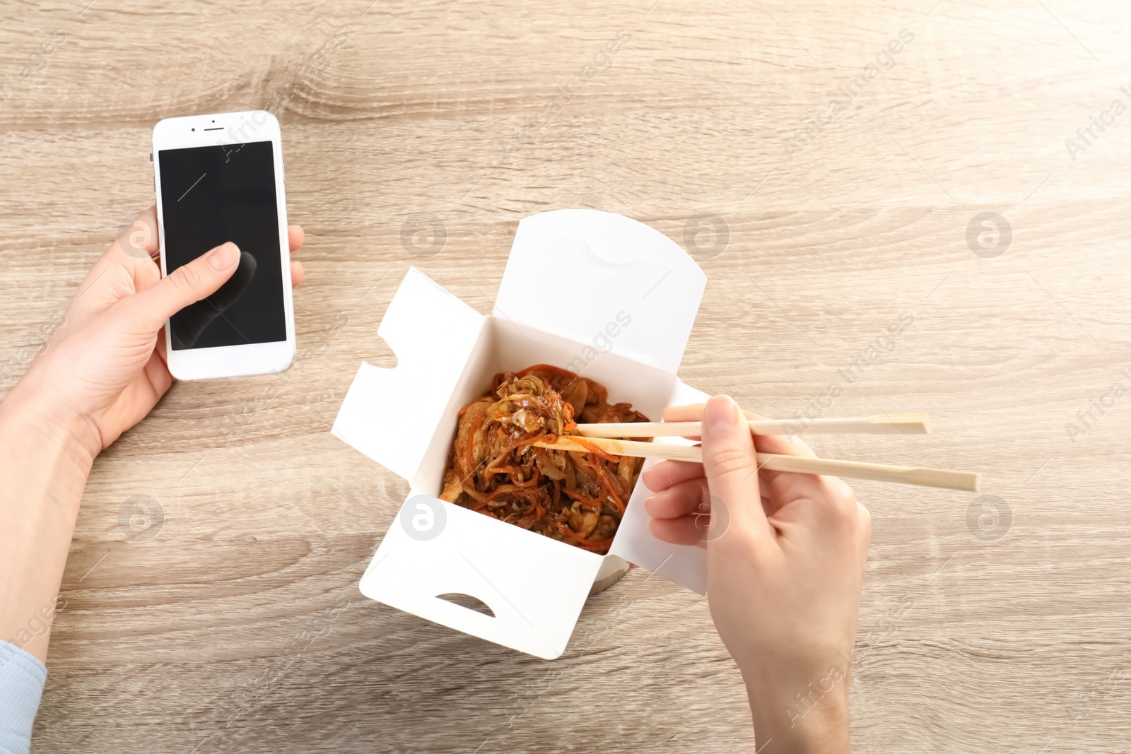 Photo of Woman holding smartphone with space for text while eating Chinese noodles at wooden table, closeup. Food delivery