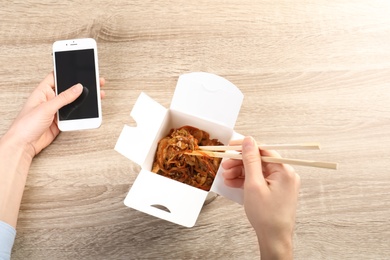 Photo of Woman holding smartphone with space for text while eating Chinese noodles at wooden table, closeup. Food delivery