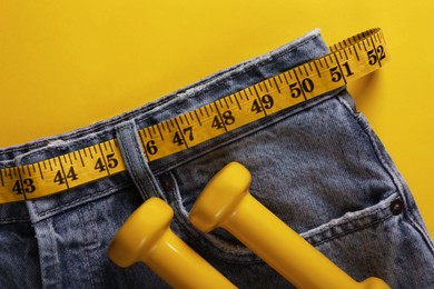 Jeans with measuring tape and dumbbells on yellow background, top view. Weight loss concept