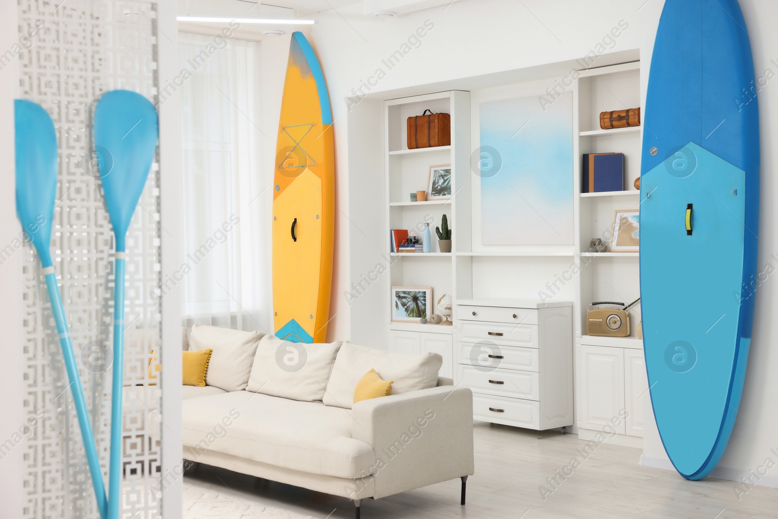 Photo of SUP boards, shelving unit with different decor elements and stylish sofa in room. Interior design