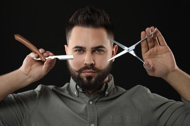 Photo of Handsome young man with mustache holding blade and scissors on black background
