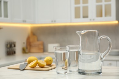 Photo of Jug, glasses with clear water and lemons on white table in kitchen