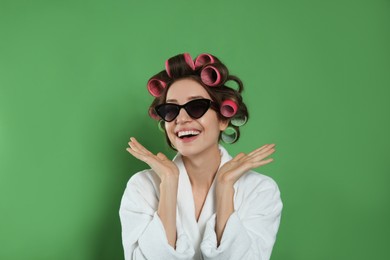 Beautiful young woman in bathrobe with hair curlers and sunglasses on green background